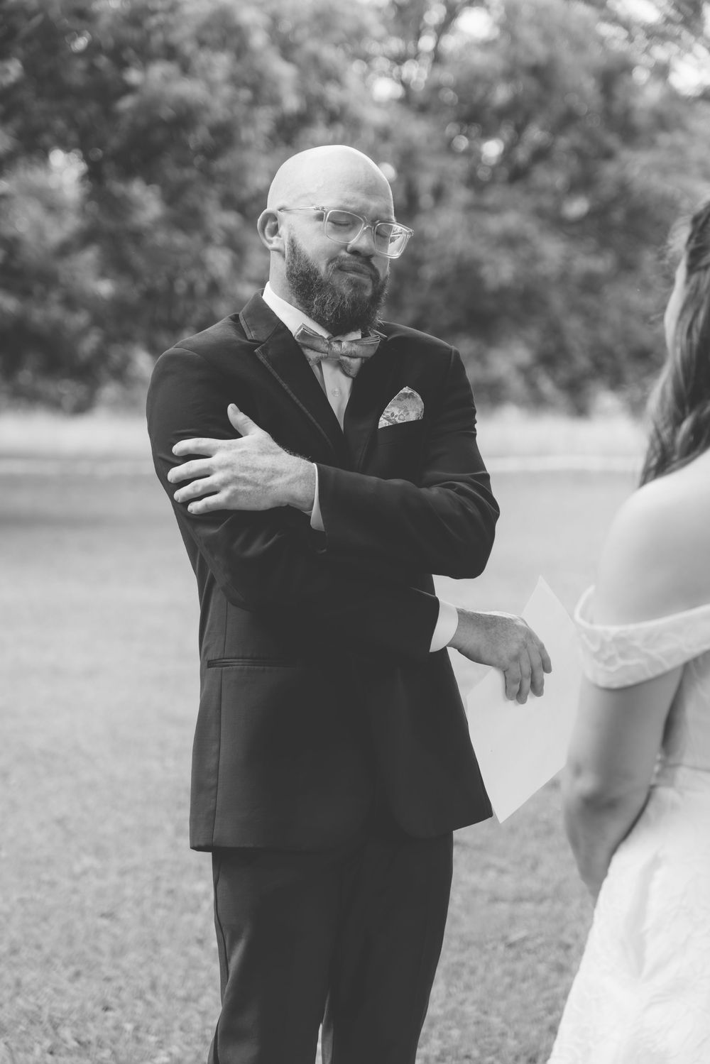 Emotional bald bearded groom in tuxedo with bow tie crossing arms during wedding vows ceremony outdoors in black and white.