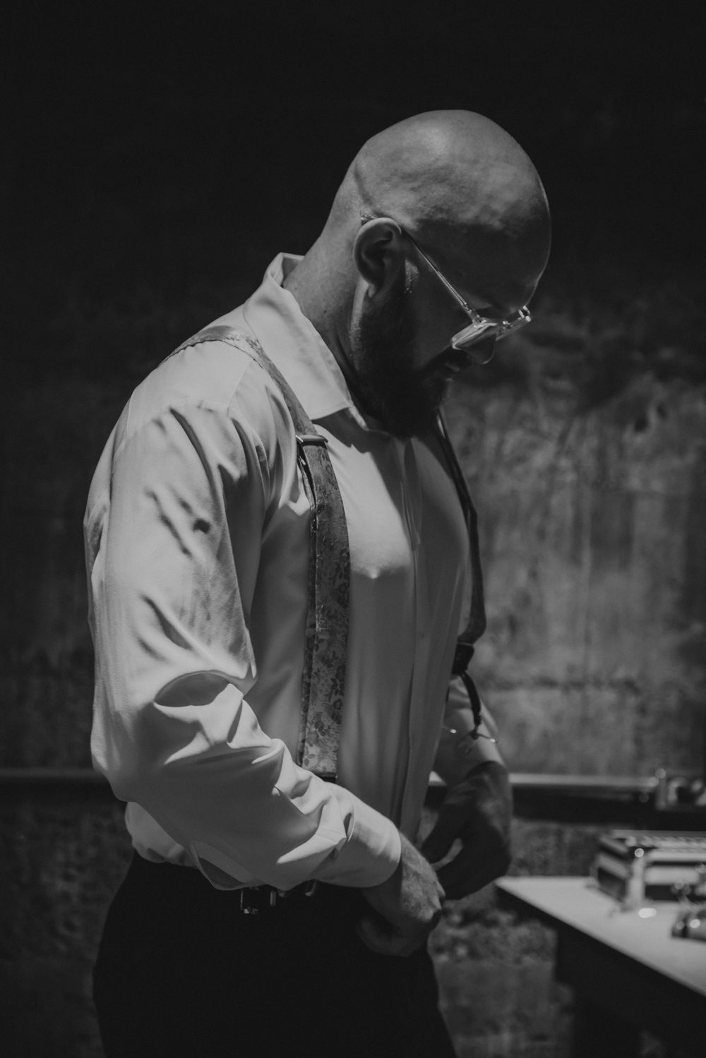 Bald bearded groom in white dress shirt and suspenders preparing for wedding ceremony in dramatic black and white photo.