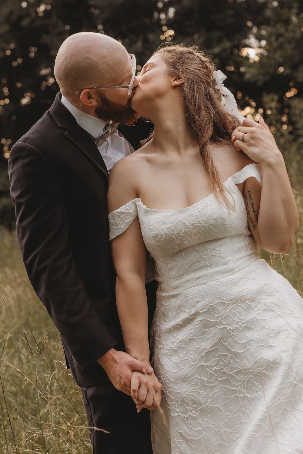 A bald groom in a dark suit kisses his bride in an off-shoulder white lace gown outdoors in a field.