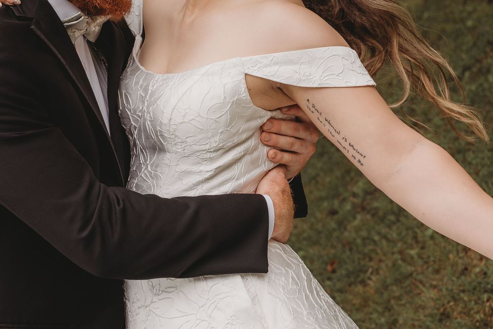 Close-up of groom holding bride in off-shoulder lace wedding dress revealing meaningful script tattoo on her arm.