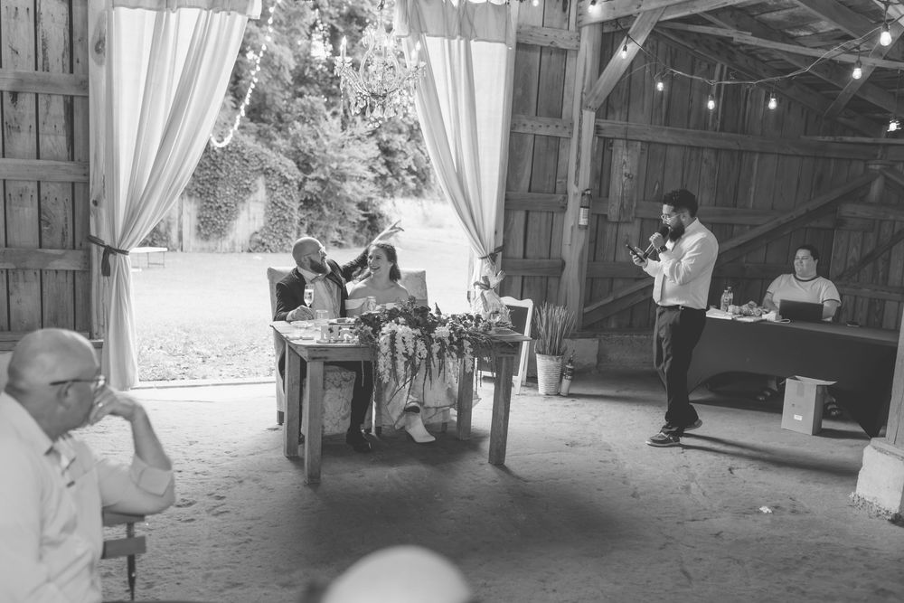 Wedding guest delivers speech at rustic barn reception while bride and groom sit at head table.