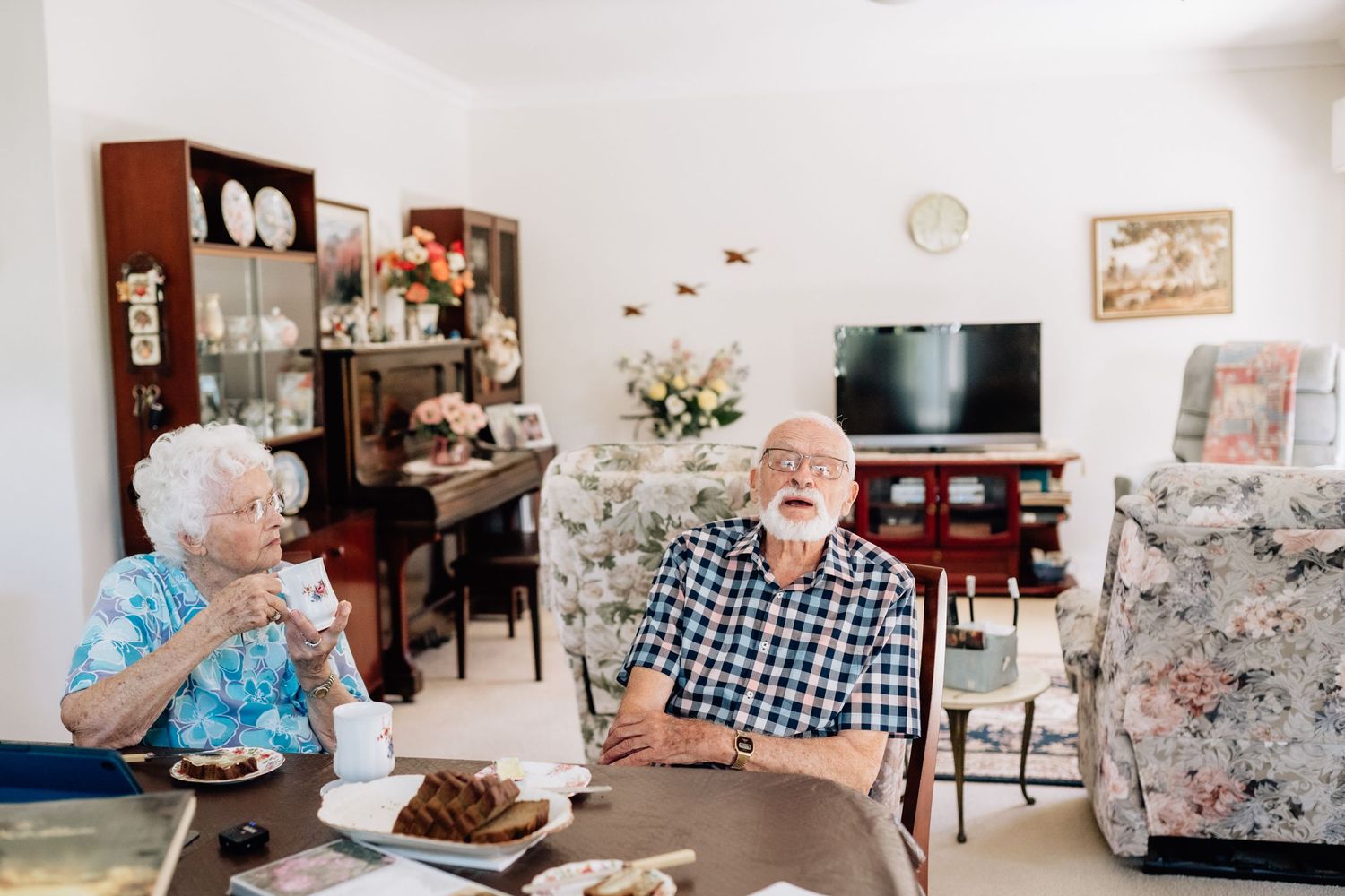 An elderly man and woman sit at a dining table with tea and cake.