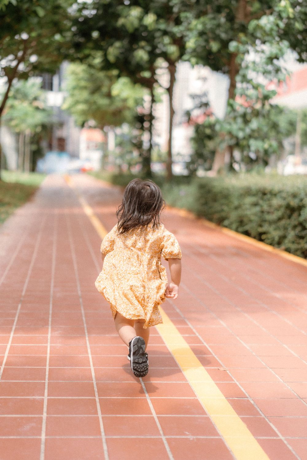 Child in yellow dress running along red brick path surrounded by greenery.