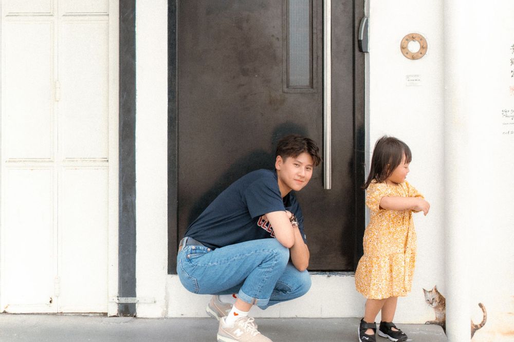 Two figures sit on a doorstep, one crouched and one standing in a yellow dress against a white wall.