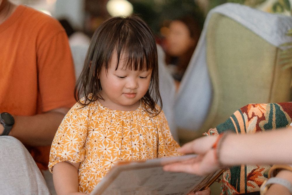 A small child in an orange floral dress looks down at a book.