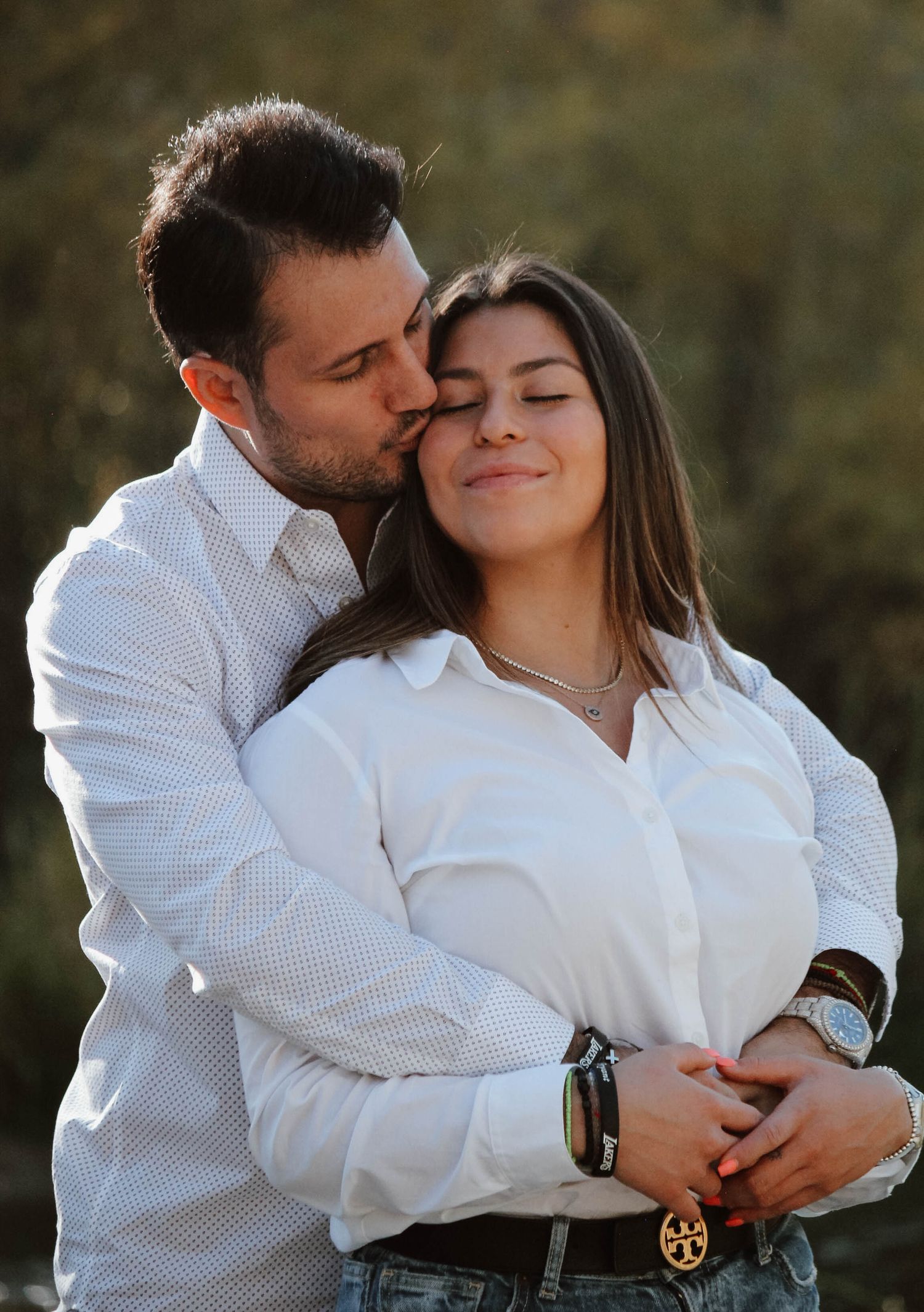 A tender embrace between a couple wearing white shirts against a natural outdoor backdrop.