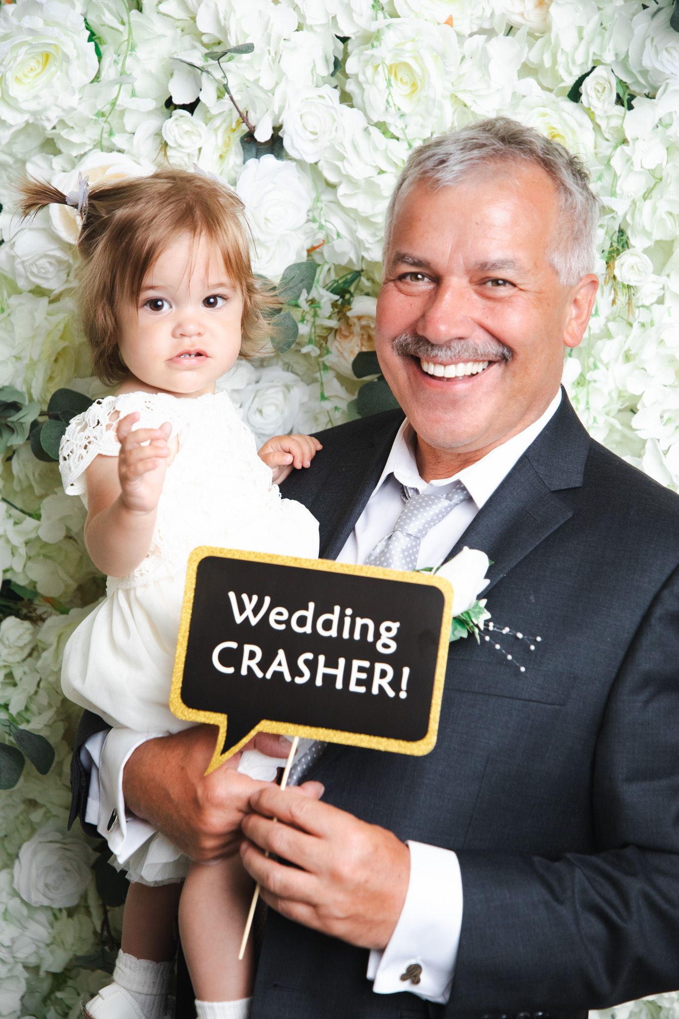 A fun photo booth moment with props showing 'Wedding Crasher' signs against a white floral backdrop.