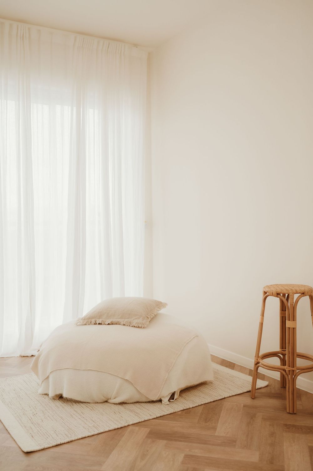 Minimalist room interior with white ottoman on cream rug, bamboo stool and sheer curtains filtering natural light.
