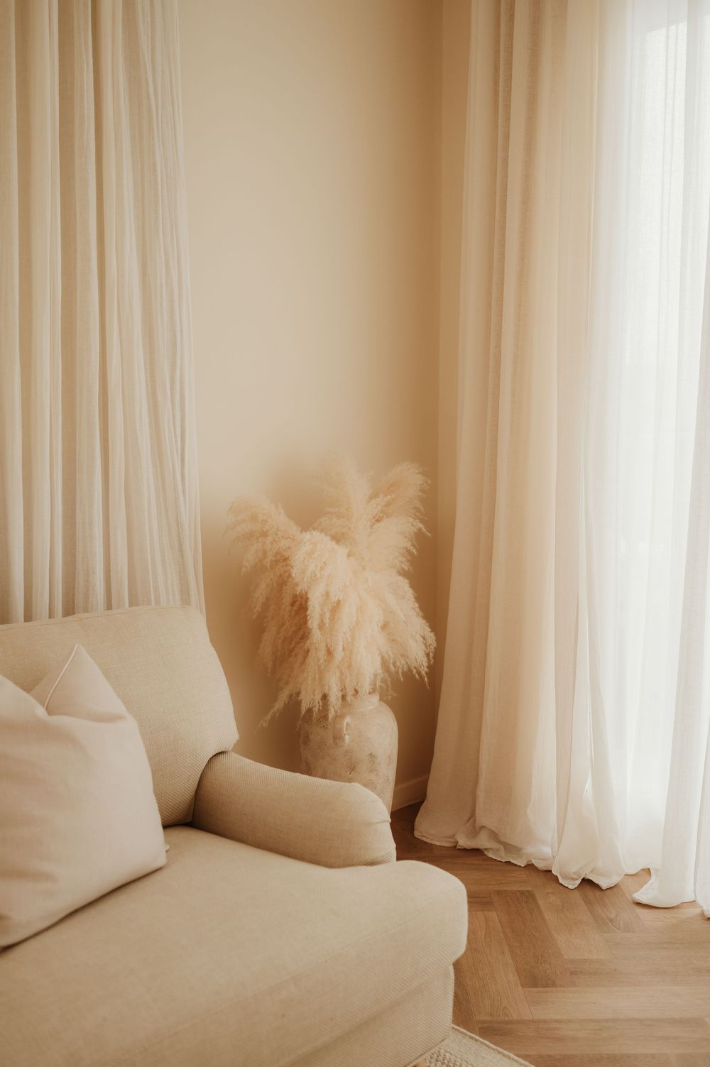 Minimalist interior setting with flowing white curtains, beige cushions, and decorative pampas grass in soft lighting.