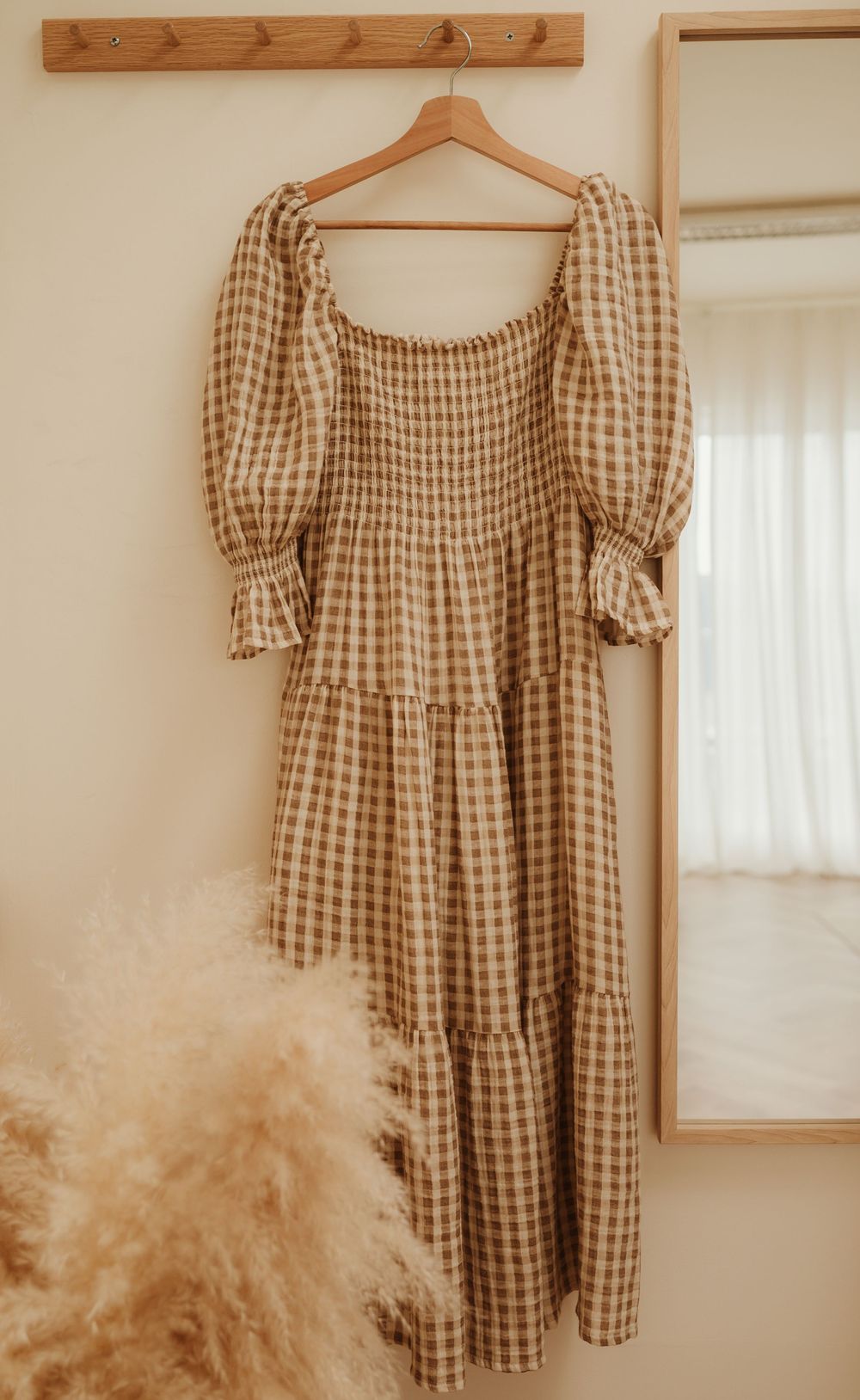 Wooden five-hook wall mounted coat rack and brown gingham puff sleeve dress displayed on a hanger in warm sunlight.
