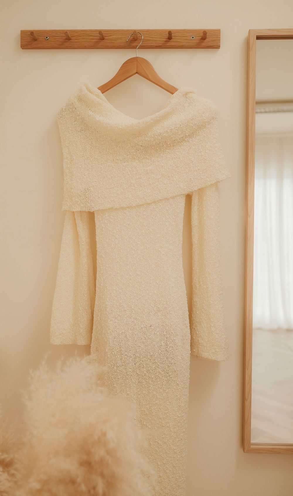 A cream colored cowl neck sweater dress hangs on a wooden hanger against a white wall next to a mirror.