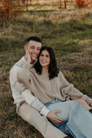 Two people in cozy knit sweaters sit close together on grass during a warm autumn sunset.