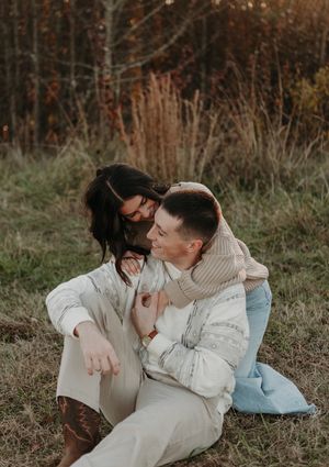 A couple wearing light colored sweaters embraces while sitting together outdoors in an autumn field setting.