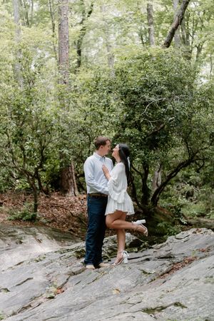 A couple embraces romantically on a rocky outcrop surrounded by lush green forest scenery.