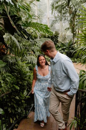 A couple walks together through a lush greenhouse conservatory filled with tropical palm plants and greenery.