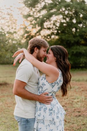 A couple in casual attire share a romantic embrace outdoors at sunset with trees in the background.