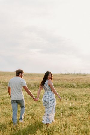 A couple walks hand in hand through a golden wheat field on a cloudy summer evening.