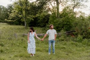 Two people hold hands while walking through a grassy meadow surrounded by trees at sunset.