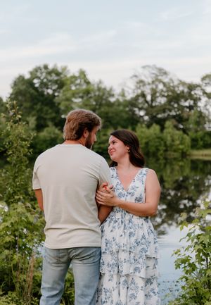 A couple stands together by a serene pond on a summer evening, with lush greenery surrounding them in a romantic setting.