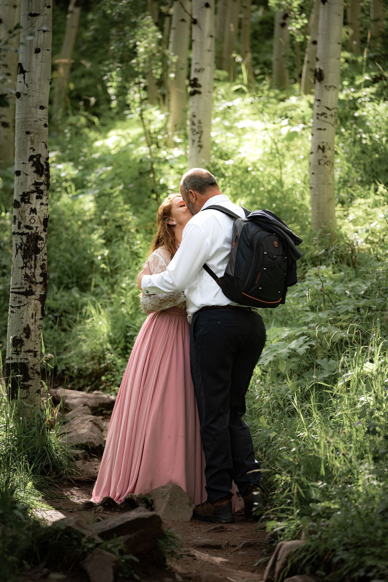 A tender embrace between two people in formal wear on a forest path surrounded by aspen trees.