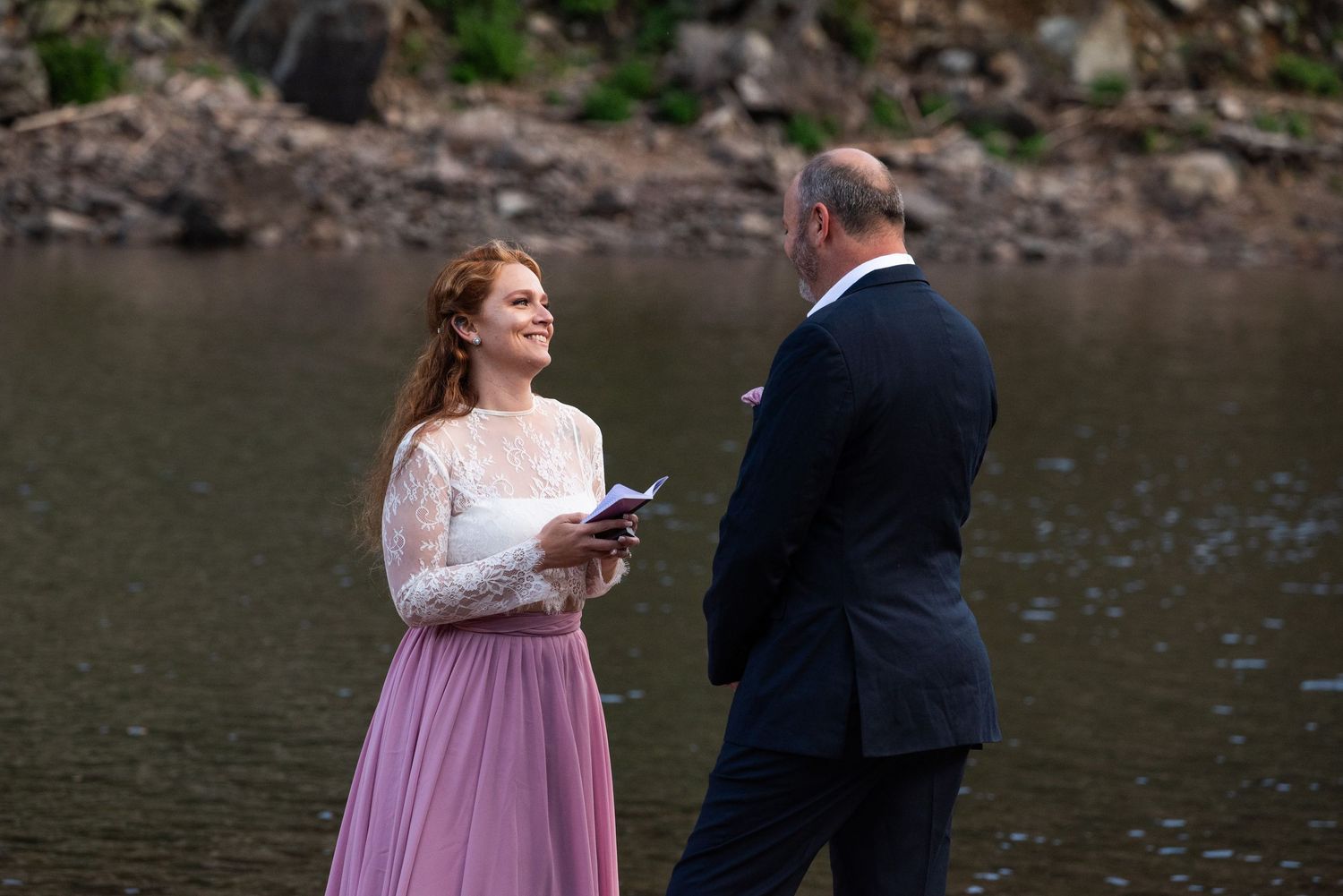 A couple in formal attire sharing a moment by a mountain lake during what appears to be a ceremony.