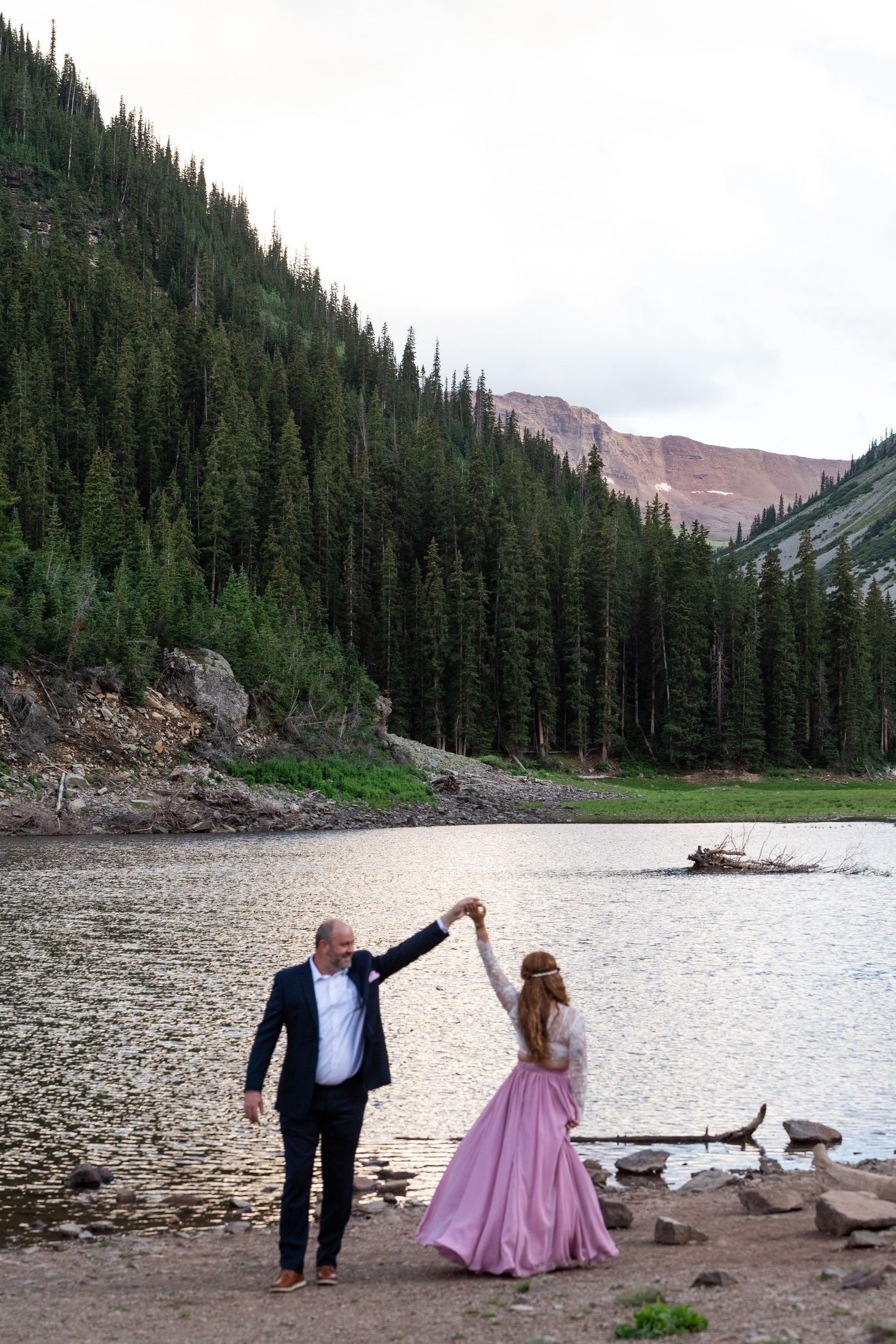 A couple in formal attire dancing by a mountain lake surrounded by evergreen forest.