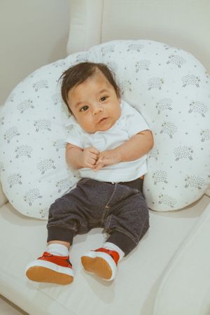 Baby wearing white top and pants rests on a patterned nursing pillow.