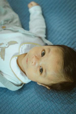 A young child wearing white lies on blue bedding looking up at the camera.