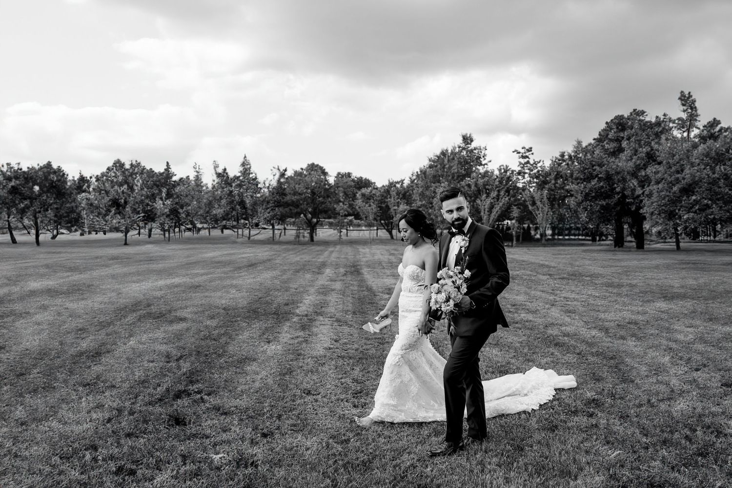 A black and white wedding photo of a couple standing in an open grassy field with trees in the background.