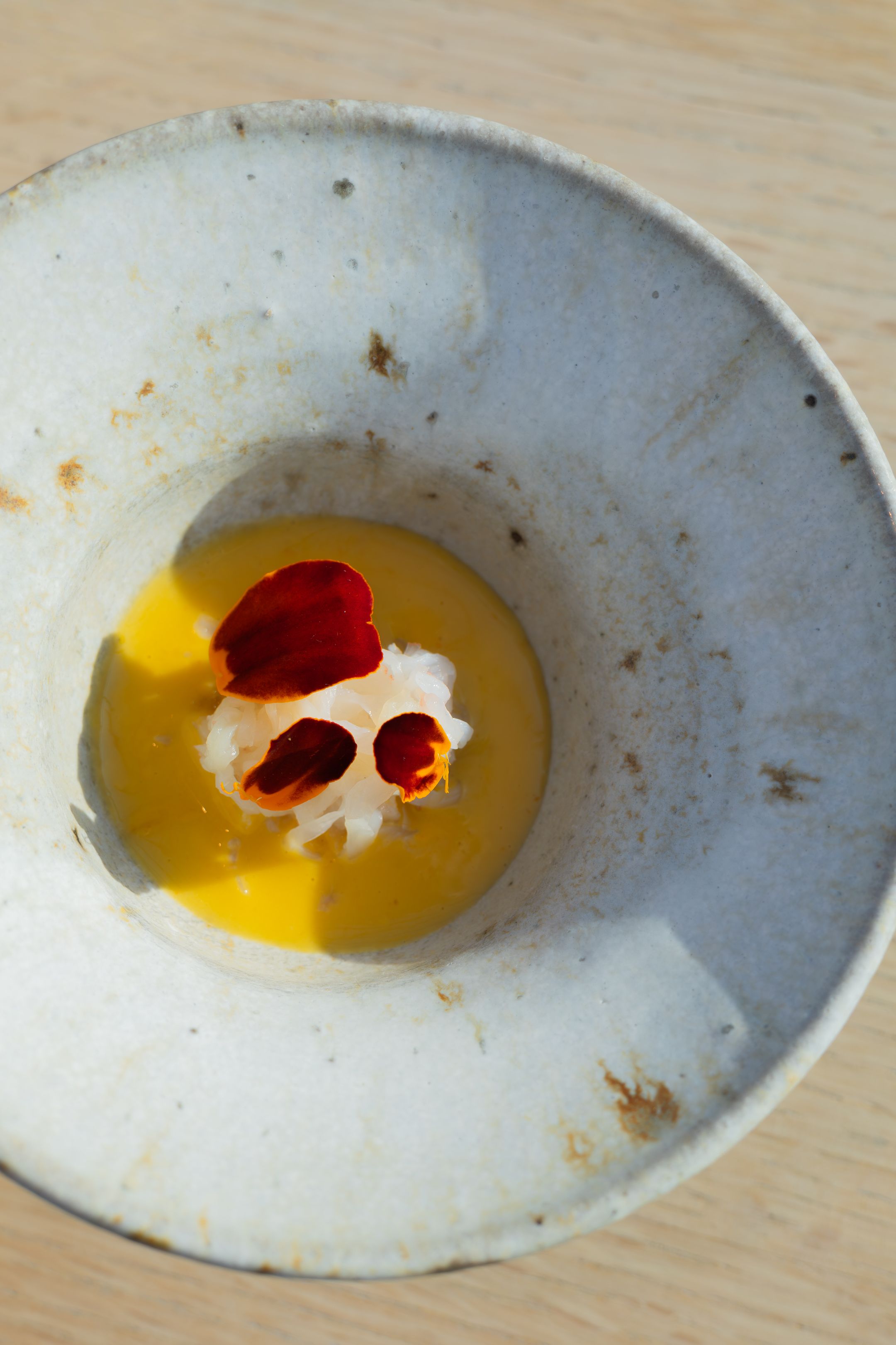 Yellow soup garnished with red petals and white cream in rustic ceramic bowl.