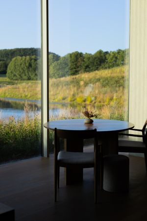Minimalist dining setup with round table against floor-to-ceiling windows overlooking a pond.