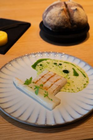 Grilled fish fillet on a white plate with green sauce and herbs, accompanied by rustic bread.