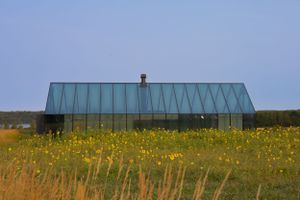 A modern glass house sits in a field of yellow wildflowers against a blue sky.