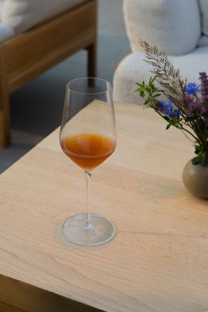 A glass of orange-hued wine sits on a wooden table with delicate wildflowers in a vase.