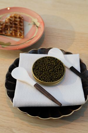 Caviar tin with serving spoons on black plate next to heart-shaped waffle.