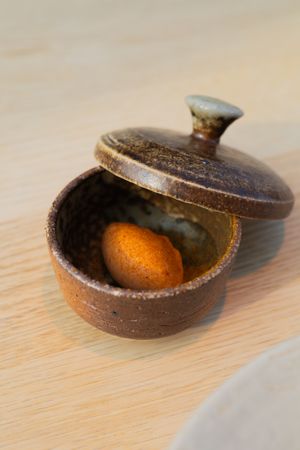 Rustic ceramic pot with lid containing a single citrus fruit on a wooden surface.
