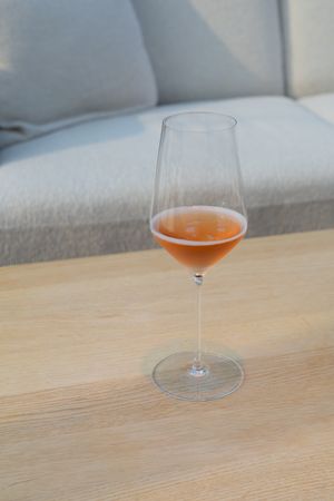 Glass of orange-tinted wine in a stemmed glass on wooden table with grey couch background.