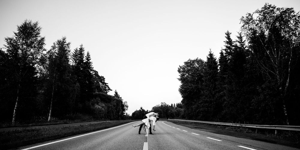 A lonely person walks down the middle of an empty tree-lined highway in black and white photography.