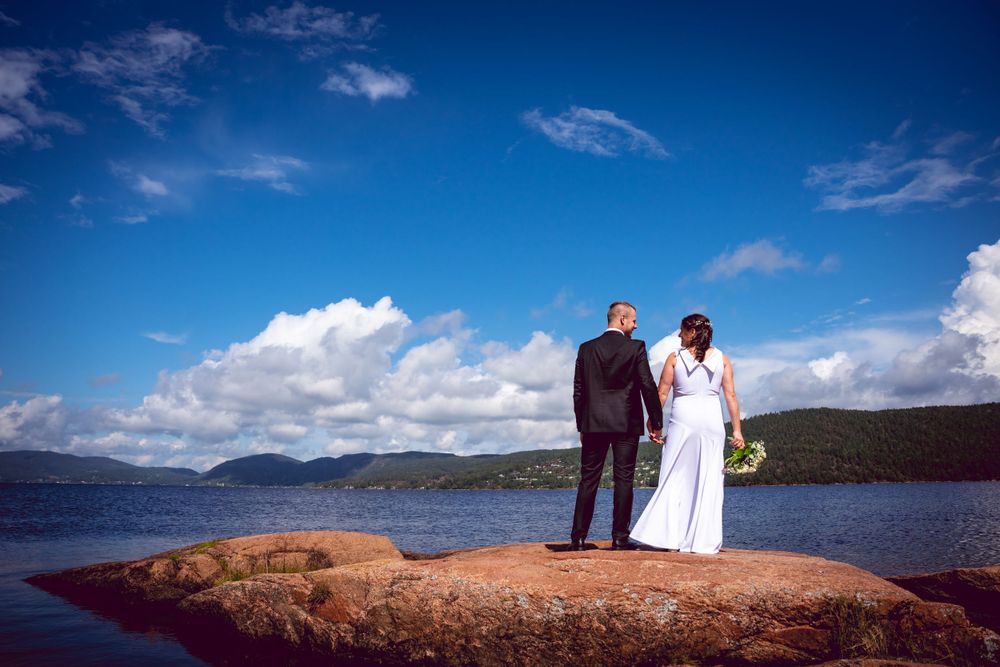 Wedding couple stands on rocky outcrop with scenic lake and mountains in background.