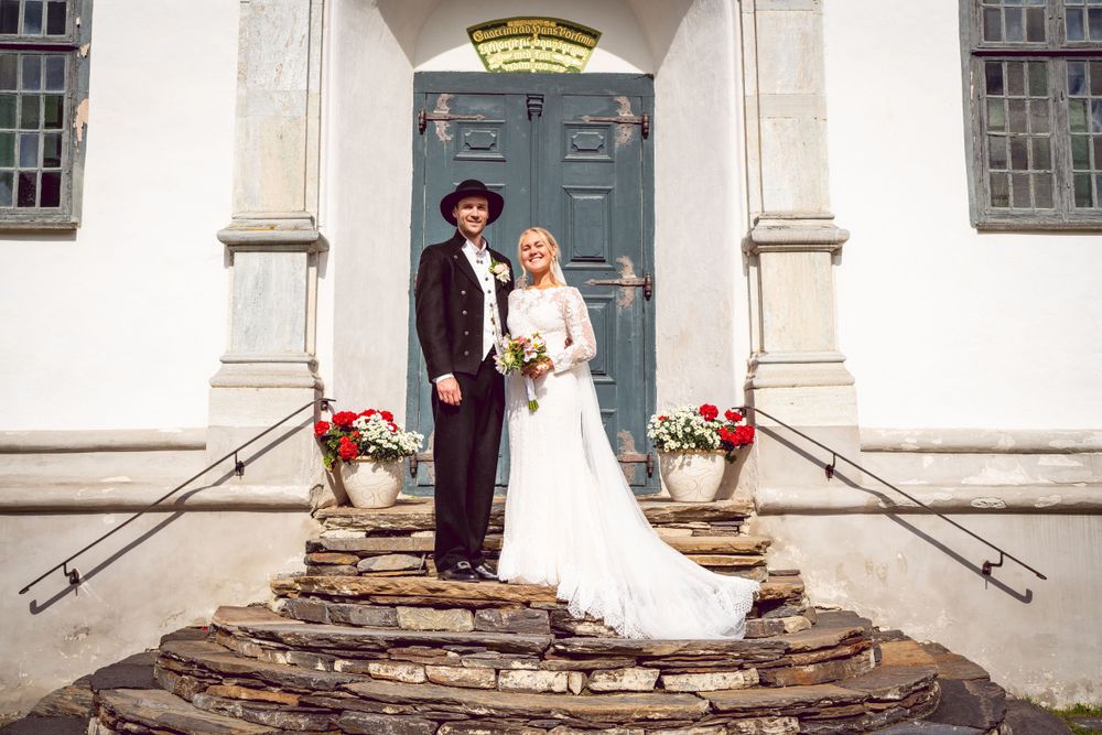 Wedding portrait on stone steps of historic white church with red floral decorations.