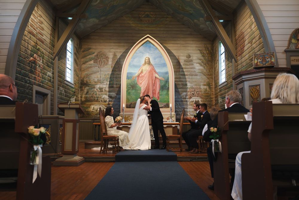 A couple shares a kiss at the altar during a church wedding ceremony with guests seated in wooden pews.