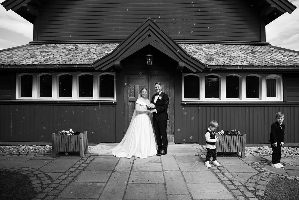 Black and white wedding photo of bride and groom posing outside a rustic barn venue with two children nearby.