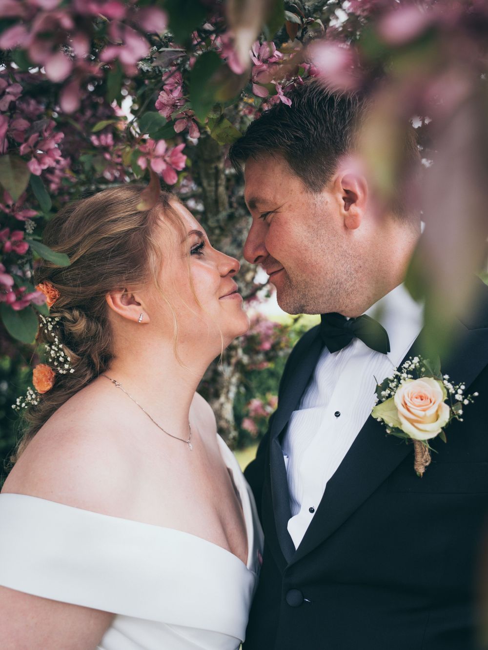 Bride and groom share a romantic moment under blooming pink flowers, groom in tuxedo with rose boutonniere.