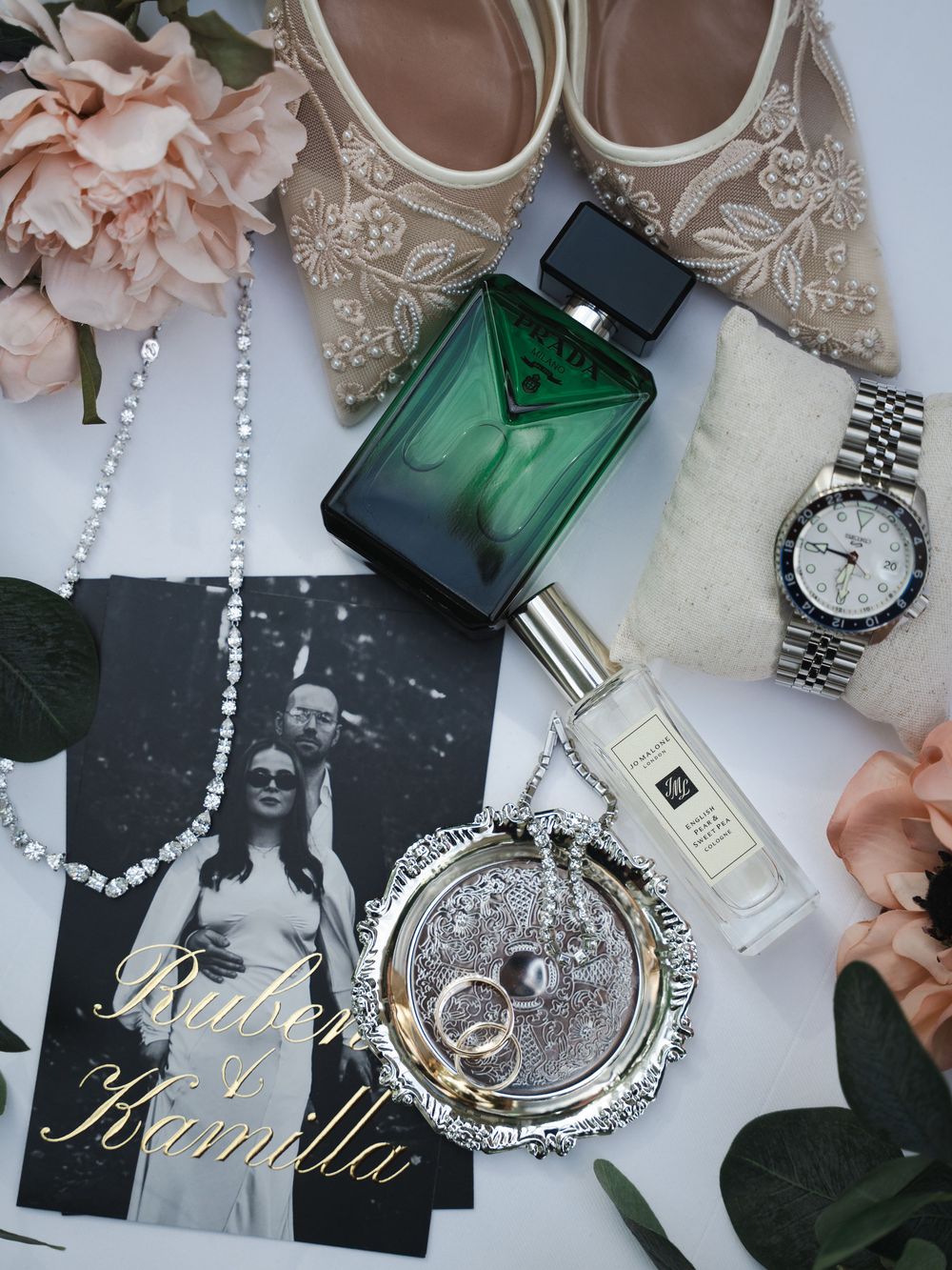 Elegant wedding flat lay with perfume, Jo Malone cologne, rings, diamond necklace, watch, heels, and personalized name card.