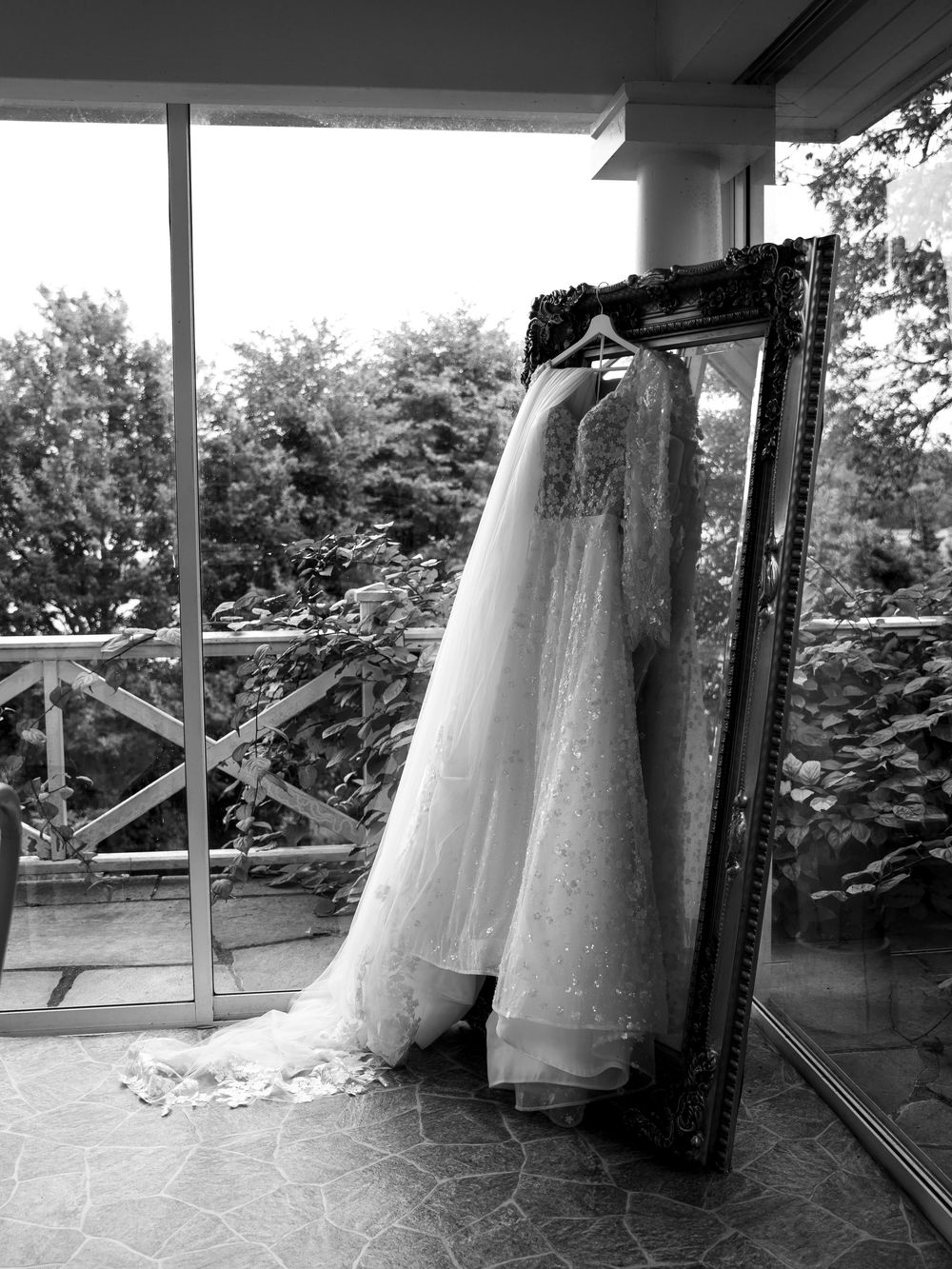 Black and white photo of an elegant lace wedding gown hanging on an ornate mirror near a window with garden views.