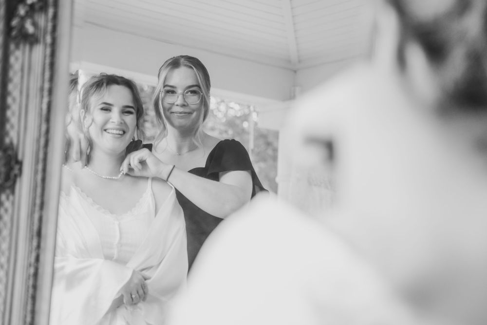 Black and white photo of two smiling women looking in a mirror during bridal preparations.