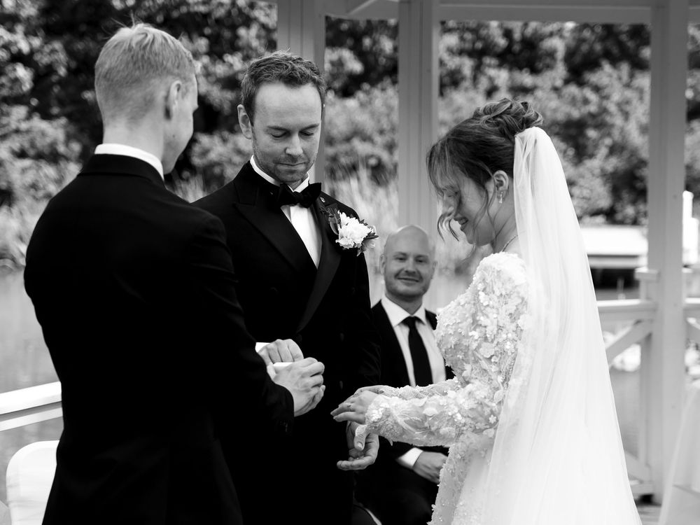 Black and white wedding ceremony photo of bride and groom exchanging rings with officiant in elegant outdoor setting.