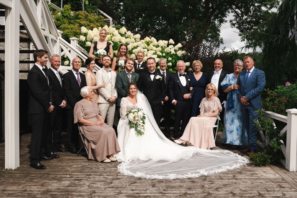 Wedding group photo with bride in white gown and veil, groom in tuxedo, surrounded by family outdoors near white stairs.