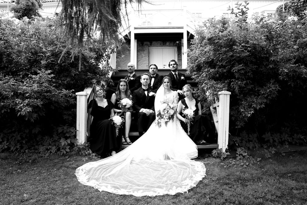 Black and white wedding photo of bride, groom, and bridal party posing outdoors near a historic home.