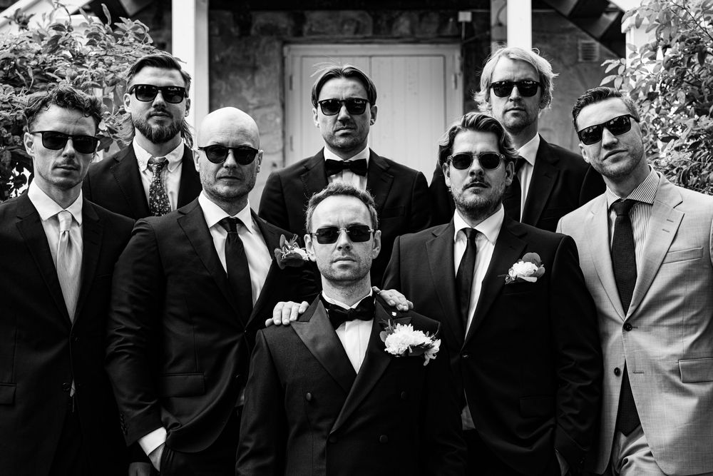 Black and white photo of a groom and eight groomsmen in suits and sunglasses posing together outdoors.